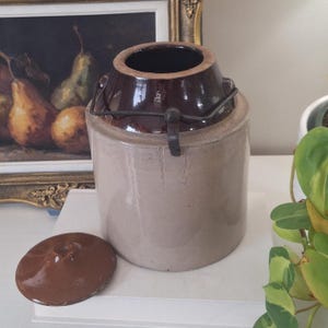Peut inclure: Un pot en grès cylindrique avec un corps beige clair et un bord émaillé brun foncé. Le pot est doté d'une anse en fil métallique fixée au col et comprend un couvercle circulaire assorti, émaillé brun foncé, posé à côté de lui.
