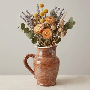 May include: A rustic brown ceramic pitcher with a speckled finish and a single handle, filled with a dried floral arrangement featuring yellow billy balls, purple lavender sprigs, orange strawflowers, poppy pods, and green eucalyptus leaves.