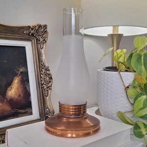 May include: A lamp component featuring a circular copper-coloured metal base with a tiered design. Attached to the base is a tall, bulbous glass chimney that transitions from a frosted, opaque white midsection to a clear, transparent top opening.