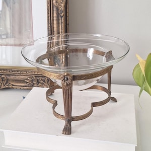 Peut inclure: Un saladier en verre transparent sur un support décoratif en laiton. Le support présente des détails ornés et une base à trois pieds. Le saladier rond repose sur le cadre en laiton, créant une pièce d'exposition élégante.