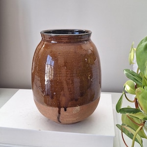 May include: A brown ceramic vase with a rounded body and a dark interior. The vase has a glossy finish with a drip of glaze near the base. The lower section of the vase is a lighter, unglazed clay colour.