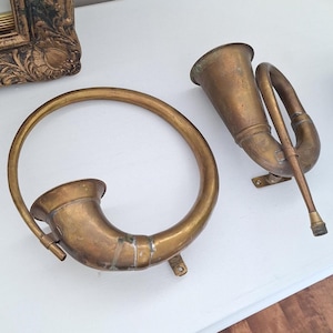 May include: Two antique brass horns. One is a circular horn with a flared bell, and the other is a trumpet-style horn with a curved body. Both have a tarnished, aged appearance, suggesting they are vintage items.