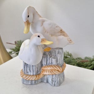 Peut inclure: Figurine en céramique représentant deux canards perchés sur un poteau en bois vieilli. Les canards sont principalement blancs avec des accents beiges et jaunes. Un détail de corde entoure le poteau. Objet décoratif.