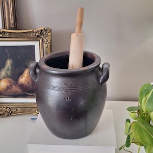 May include: A dark brown ceramic crock with two side handles, featuring the embossed text HOGANAS and 5L on the front. A light-coloured wooden rolling pin is placed vertically inside the opening of the crock, which has a smooth, matte finish.