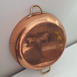 Peut inclure: Une poêle ronde en cuivre présentant une surface métallique polie avec des rayures visibles et deux poignées en laiton fixées sur les côtés opposés. La poêle a une finition réfléchissante et un design incurvé peu profond avec un bord arrondi.