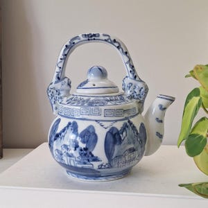 May include: A white ceramic teapot featuring blue hand-painted landscape illustrations of mountains and buildings. The teapot has a curved handle, a lid with a small knob, and a spout. A geometric Greek key pattern encircles the upper body.