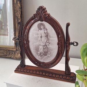 May include: A dark brown wooden oval swivel picture frame with ornate carved detailing on the base and frame edges. The frame holds a black and white photograph of a person wearing a wide-brimmed hat and a dress, holding a bouquet of flowers.