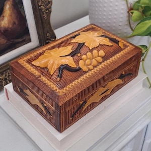 May include: A rectangular wooden box featuring a dark brown textured finish with carved light brown grape leaves and clusters of grapes on the lid and sides. The box has a decorative zigzag border pattern along the edges of the lid.