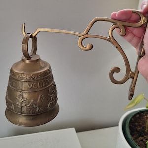 Peut inclure: Une cloche murale en laiton avec un support métallique incurvé et orné. Le corps de la cloche présente des figures animales en relief et l'inscription latine VOCEM MEAM. La cloche est suspendue à un anneau circulaire fixé au bras décoratif.