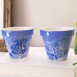 May include: Two ceramic flower pots with a tapered shape and a light blue rim. The exterior features a blue and white transferware print depicting a snowy winter landscape with houses and trees. The right pot includes the text Currier & Ives.