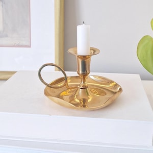 Peut inclure: Un bougeoir doré avec une bougie blanche. Le bougeoir a une base ronde avec une poignée et une coupelle pour tenir la bougie. La bougie est blanche et non allumée. Le bougeoir est sur une surface blanche.