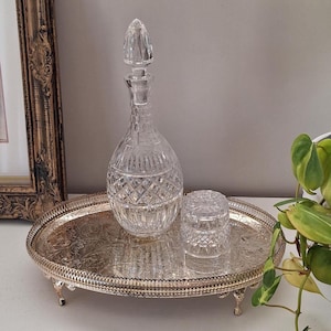 Peut inclure: Une carafe en verre transparent avec son bouchon assorti et un petit pot en verre, tous deux ornés d'un motif taillé en diamant, posés sur un plateau ovale en métal argenté avec une surface gaufrée, un bord perlé et quatre petits pieds.