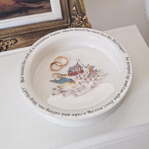May include: A round white ceramic trinket dish featuring a colourful illustration of a rabbit in a blue jacket running from a person with a rake near a greenhouse. The rim is inscribed with the text: But round the end of a cucumber frame, whom should he meet but Mr. McGregor! he jumped up and ran after Peter, waving a rake and calling out, 'Stop thief!' Two gold-toned rings rest inside the dish.
