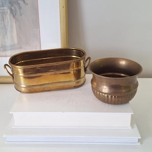May include: Two brass planters are displayed on a stack of white books. One is an oval container with handles, the other a small, round pot with vertical ridges. The planters have a warm, golden colour.
