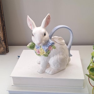 May include: A white ceramic rabbit teapot with a blue handle and bow. The rabbit features pink-lined ears and a floral garland. A delightful Easter-themed decorative item for the home, perfect for serving tea.