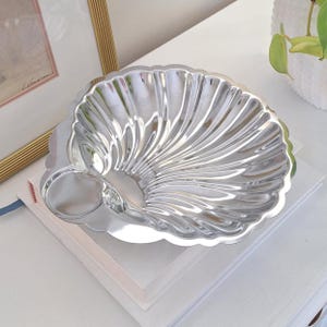 May include: A silver-coloured, shell-shaped dish with a scalloped edge. The dish has a textured, fan-like design radiating from the centre. It includes a small, round indentation on one side. The dish is sitting on a stack of white books.