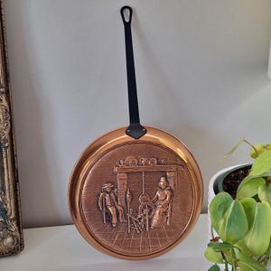 May include: A round copper wall plaque featuring a relief scene of two figures sitting by a fireplace with a spinning wheel. The plaque has a long, slender black metal handle extending from the top, designed for hanging on a vertical surface.