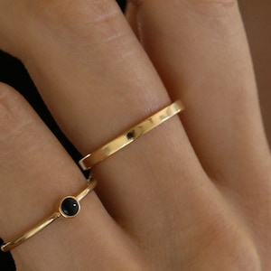 Peut inclure: Deux bagues en or sont présentées sur le doigt d'une personne. L'une des bagues comporte une petite pierre noire ronde. L'autre bague est une fine bande simple. Les bagues sont sur un fond flou.