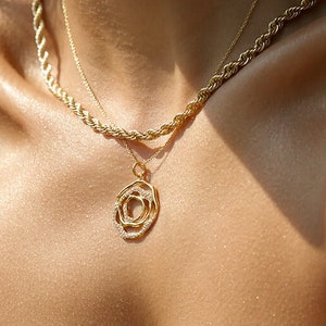 May include: A gold chain necklace with a twisted, circular pendant. The necklace is layered over a thinner gold chain.