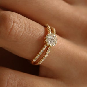 May include: A gold-colored ring with a double band design. The ring features a central, round, clear gemstone surrounded by smaller, clear gemstones. The bands are adorned with small, clear gemstones, creating a sparkling effect. The ring is on a person's finger.
