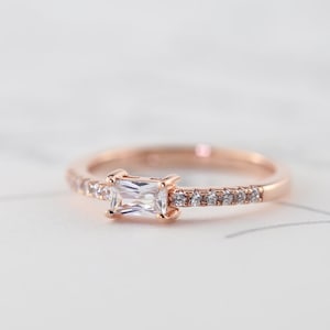 May include: Rose gold ring with a rectangular, clear gemstone at its centre. The band is embellished with small, clear gemstones. The ring is set against a soft, white background, accentuating its elegant design.