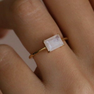 Peut inclure: Une bague en or avec une pierre de lune rectangulaire. La bague est portée sur un doigt.
