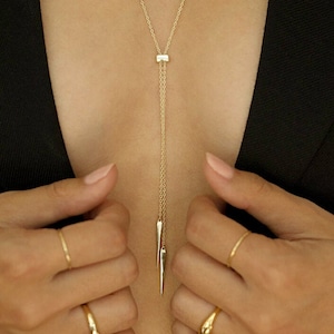 Könnte beinhalten: Eine goldene Kettenkette mit einem kleinen quadratischen Anhänger und zwei spitzen Enden. Die Kette wird von einer Person mit den Händen vor sich getragen.