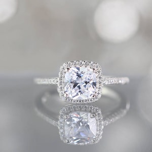 May include: A silver ring with a large, square-cut, clear gemstone surrounded by smaller stones. The ring is set on a reflective surface, creating a mirrored effect. The band is also adorned with small stones.