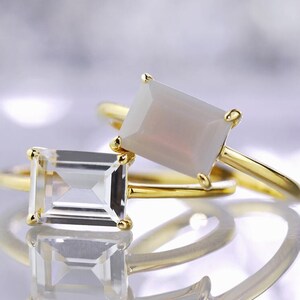 May include: Two gold rings with rectangular gemstones. The top ring has a white opal gemstone and the bottom ring has a clear quartz gemstone.