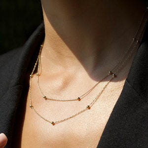 May include: A delicate gold necklace with small beads, layered on a model wearing a black blazer.