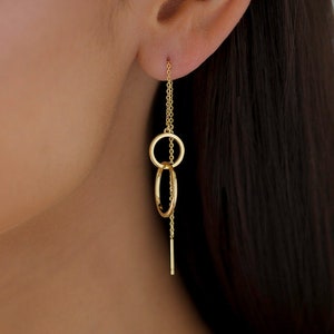 May include: Gold dangle earrings featuring two interlocking circles and a chain. The earrings have a simple, minimalist design.