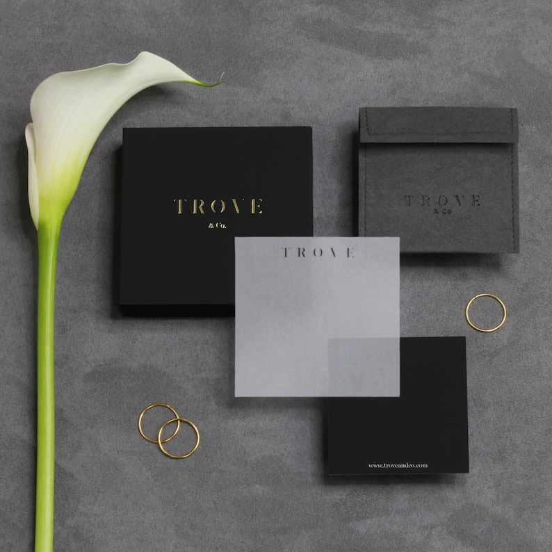 May include: Black and white packaging with gold lettering for a jewelry brand called "Trove & Co.". The packaging includes a box, a pouch, and a small card with the brand name and website address.