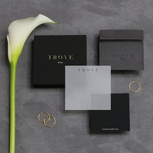 K&ouml;nnte beinhalten: Zwei schwarze Boxen mit dem Text "TROVE & Co." in goldenen Buchstaben. Eine wei&szlig;e Karte mit dem Text "TROVE" in goldenen Buchstaben liegt auf einer schwarzen Box. Eine wei&szlig;e Calla-Lilie befindet sich im Hintergrund. Der Text "www.troveandco.com" befindet sich auf der Unterseite der schwarzen Box.