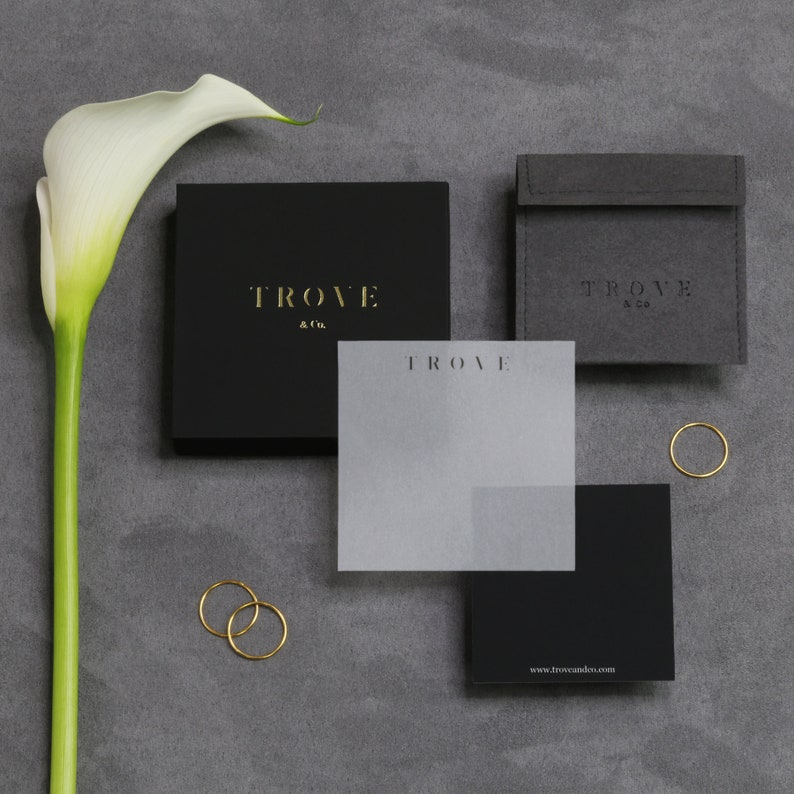 K&ouml;nnte beinhalten: Zwei schwarze Boxen mit dem Text "TROVE & Co." in goldenen Buchstaben. Eine wei&szlig;e Karte mit dem Text "TROVE" in goldenen Buchstaben liegt auf einer schwarzen Box. Eine wei&szlig;e Calla-Lilie befindet sich im Hintergrund. Der Text "www.troveandco.com" befindet sich auf der Unterseite der schwarzen Box.