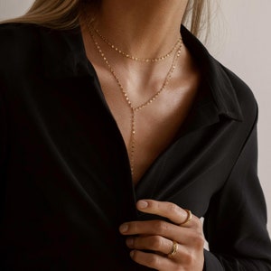 K&ouml;nnte beinhalten: Eine mehrreihige goldene Halskette mit einer langen Anh&auml;ngerkette. Die Kette wird von einer Person mit einem schwarzen Oberteil getragen.