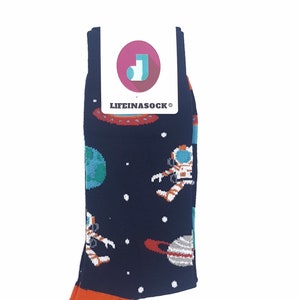 Könnte beinhalten: Marineblaue Socken mit orangefarbener Spitze und einem Weltraum-Design mit Astronauten, Planeten und Raumschiffen. Auf dem Etikett steht "LIFEINASOCK®". Das Design enthält weiße, blaue und orangefarbene Elemente.
