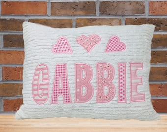 May include: White plush pillow with pink fabric appliques in the shape of hearts and the name "GABBIE" in pink letters.