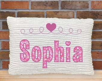 May include: A rectangular white pillow with a textured surface. The name "Sophia" is embroidered in pink, floral-patterned letters. Above the name is a pink heart and decorative swirls. The pillow is on a wooden surface.