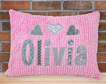May include: Pink pillow with the name Olivia embroidered in gray fabric with a patchwork design. The pillow is decorated with three gray hearts.