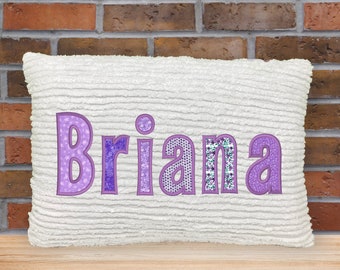 May include: A white textured pillow with the name "Briana" embroidered in purple. The letters have various patterns, including polka dots and floral designs. The pillow is rectangular and sits on a wooden surface.