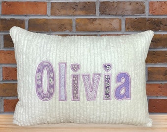May include: A white plush pillow with the name "Olivia" embroidered in purple fabric with a floral pattern.