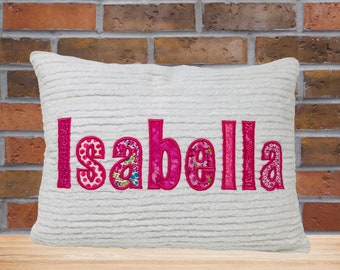 May include: White pillow with the name "Isabella" spelled out in pink fabric letters. The letters are made of different patterned fabrics.