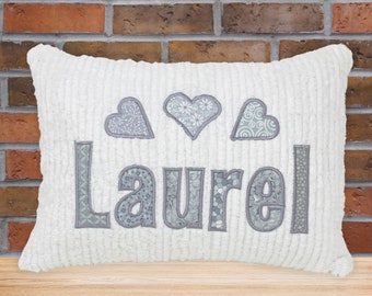 May include: White plush pillow with the name "Laurel" embroidered in gray fabric with a patchwork design. Three gray hearts with patchwork designs are above the name.