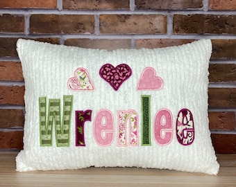 May include: White pillow with the name "Wrenlee" embroidered in pink, green, and burgundy fabric. The name is surrounded by three hearts, one pink, one burgundy, and one with a floral pattern.