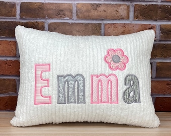 May include: White pillow with a pink and gray embroidered name "Emma" and a pink flower with a gray center.
