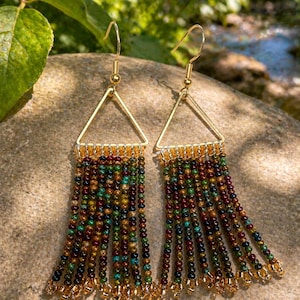 Pode incluir: Brincos triangulares dourados com franjas de contas. Os brincos apresentam uma parte superior triangular dourada com fios de contas coloridas em tons de castanho, verde e azul. Para orelhas furadas.