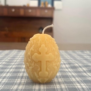 May include: A yellow egg-shaped candle with a carved cross design. The candle has a textured surface and a white wick. The candle is sitting on a blue and white checkered surface.