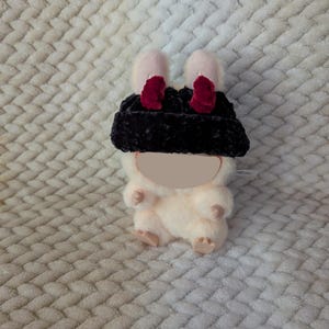 May include: A plush toy in the shape of a small animal, wearing a black hat with pink ears and red accents. The toy is cream-colored with small arms and legs. The background is a textured, off-white fabric.
