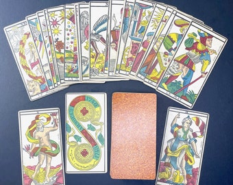 May include: A vintage deck of tarot cards with colorful illustrations and gold accents. The cards feature images of people, animals, and symbols. The cards are laid out in a fan shape on a blue surface. The cards include the names "Le Mat", "L'Empereur", "Le Monde", "L'Amoureux", "Jupiter", and "Junon".