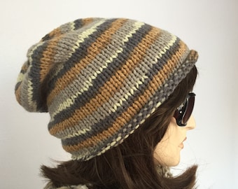 Può includere: Un cappello beanie soffice con un motivo a righe in marrone, grigio e crema. Il cappello è realizzato in tessuto a maglia e ha un bordo ripiegato.